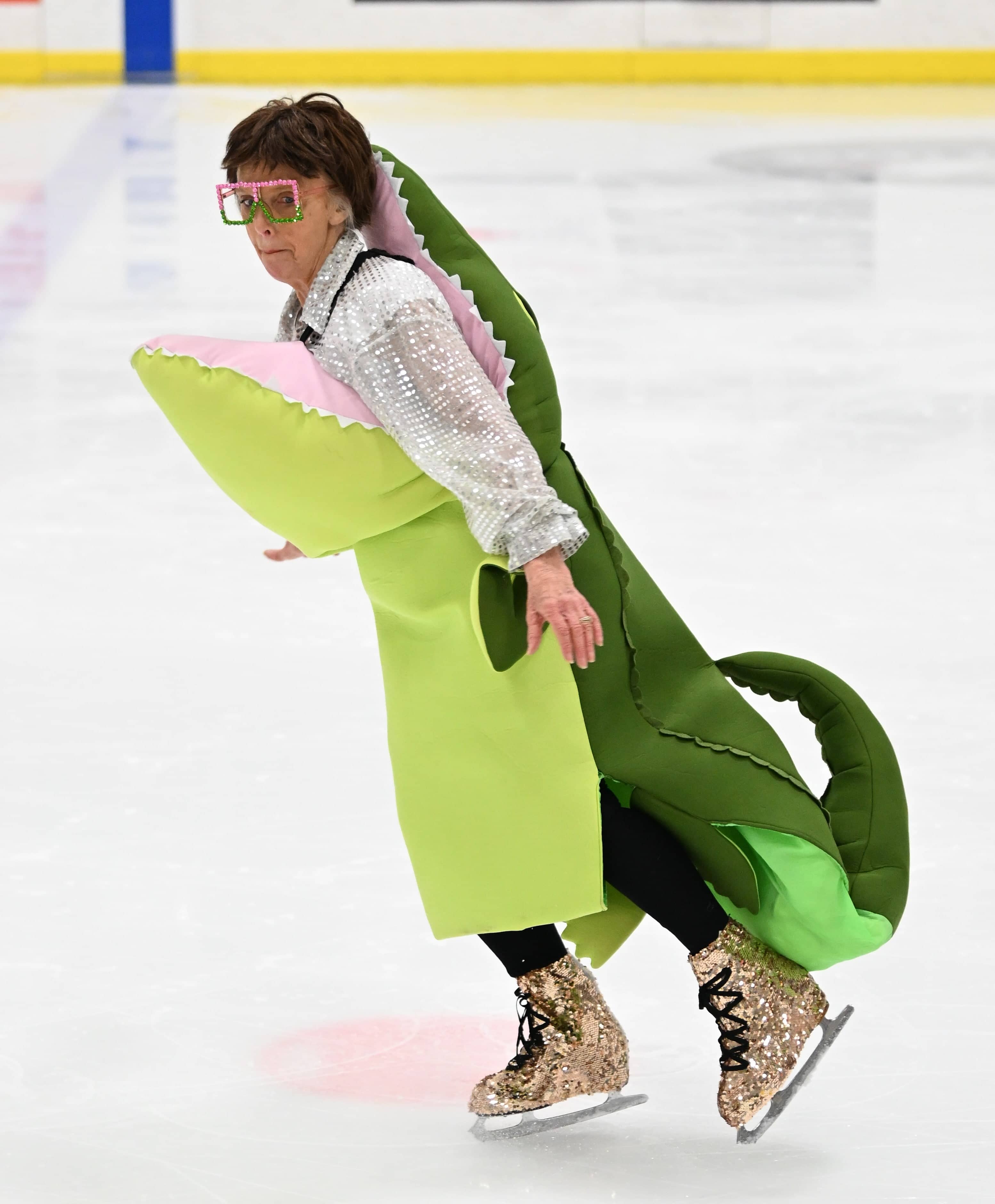 A woman with short brown hair wearing a large comedic alligator costume, large colorful glasses and gold sparkly skates